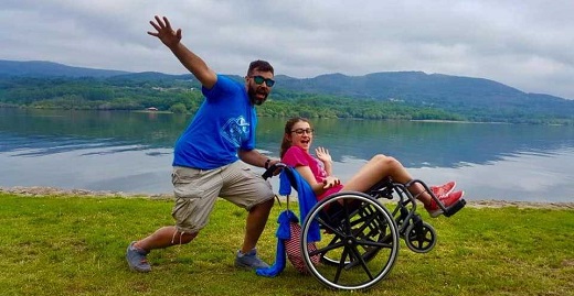 monitor sonriendo con chica en silla de ruedas frente al lago en los campamentos inclusivos