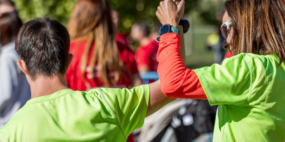 Persona participante en una actividad al aire libre levanta el brazo junto a otra persona, ambas vestidas con camisetas deportivas de color verde fluorescente. Al fondo aparecen varias personas desenfocadas en un entorno exterior con vegetación y luz diurna.