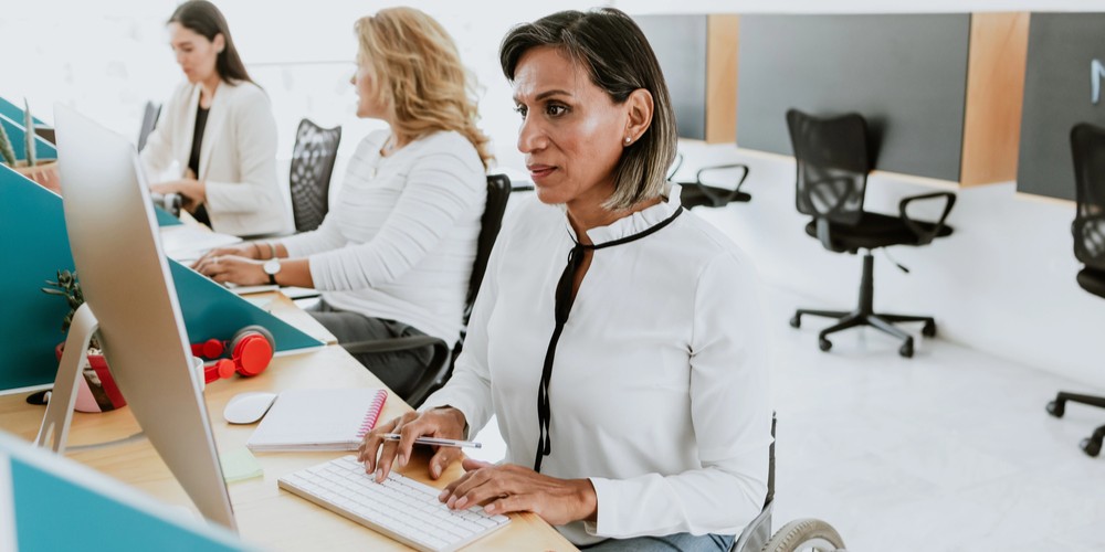 Mujer con silla de ruedas trabajando en su escritorio, al lado de dos compañeras del trabajo que están en sus respectivos sitios.