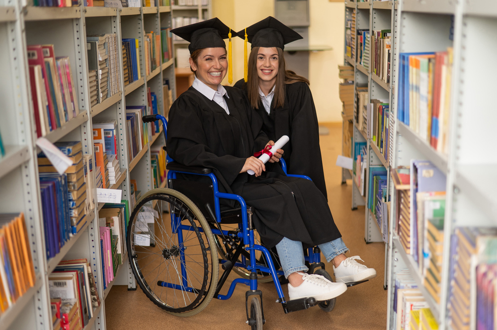 Persona usuaria de silla de ruedas y otra persona con toga y birrete posan entre estanterías de una biblioteca universitaria. La persona sentada sostiene un diploma enrollado, mientras ambas aparecen en un entorno académico rodeado de libros. La imagen representa la inclusión y el acceso a la educación superior.
