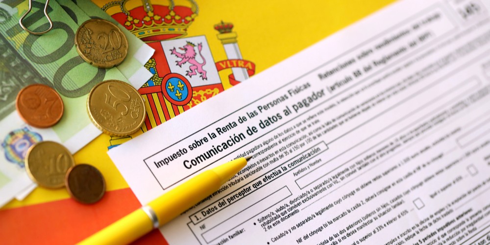 imagen del modelo 145 del IRPF con un bolígrafo al lado, también se observan billetes de 100 euros y monedas, de fondo está una bandera de España