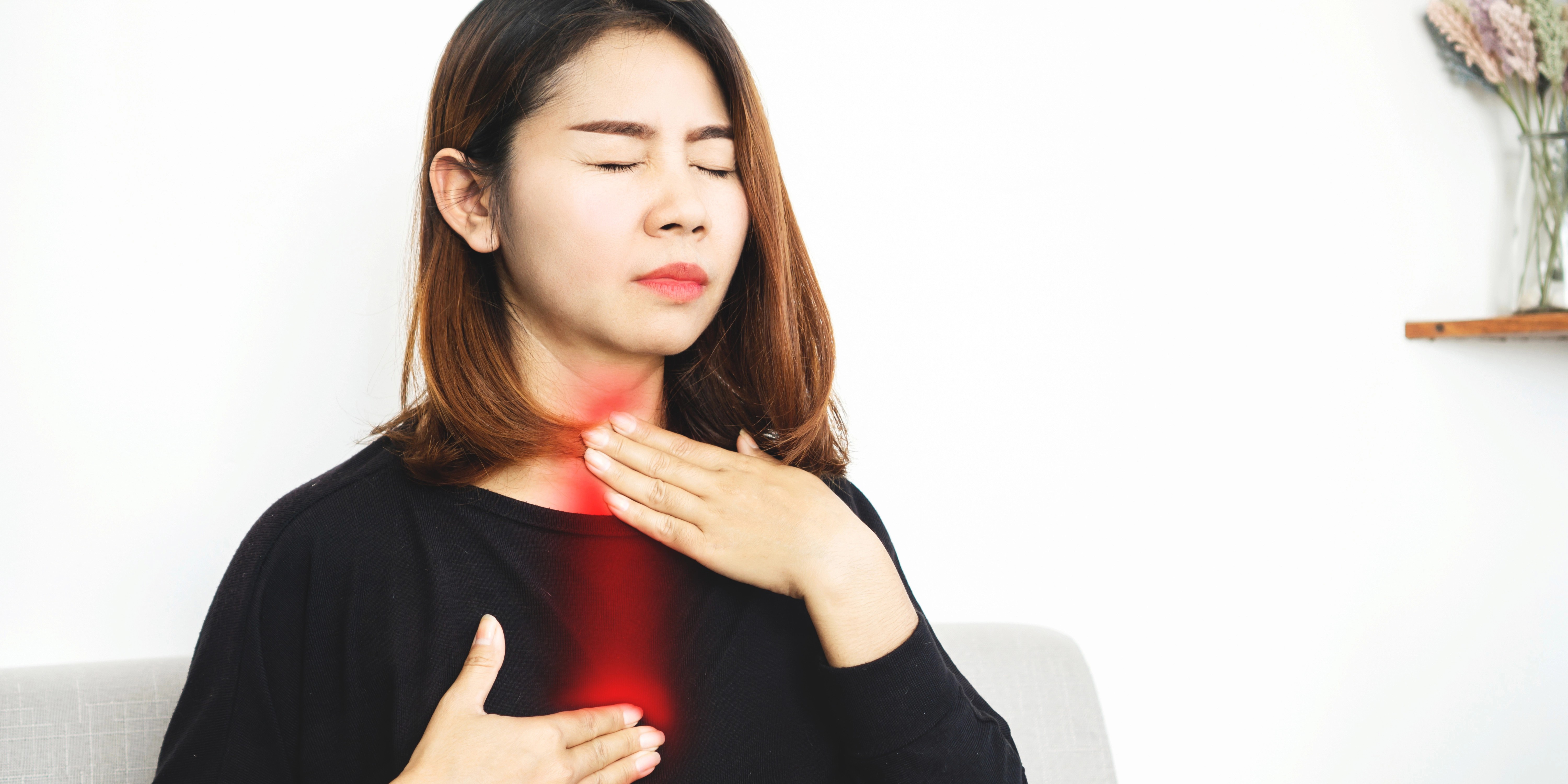 Alt texto: Persona sentada en un entorno interior con una mano sobre la garganta y otra sobre el pecho. Un efecto luminoso rojo recorre la garganta y la zona central del tórax, representando dolor, irritación o reflujo ácido. El fondo es claro y desenfocado, con una estantería y un jarrón con flores al fondo.