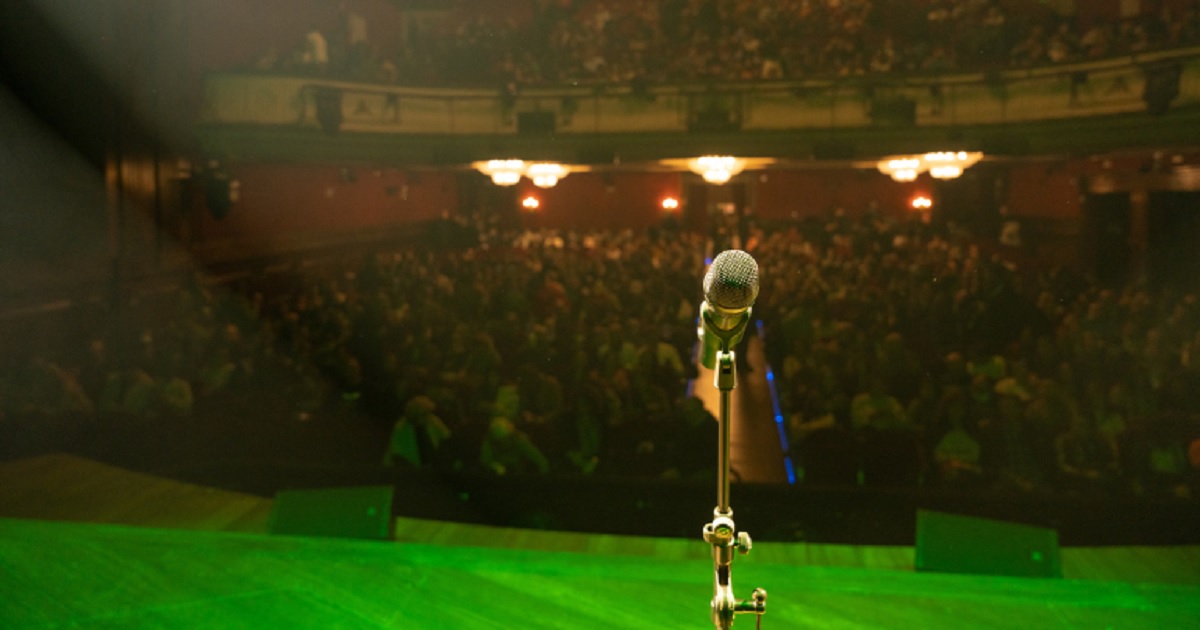 Un micrófono en un escenario y frente a un teatro lleno de gente (Fuente: Plena Inclusión Madrid)