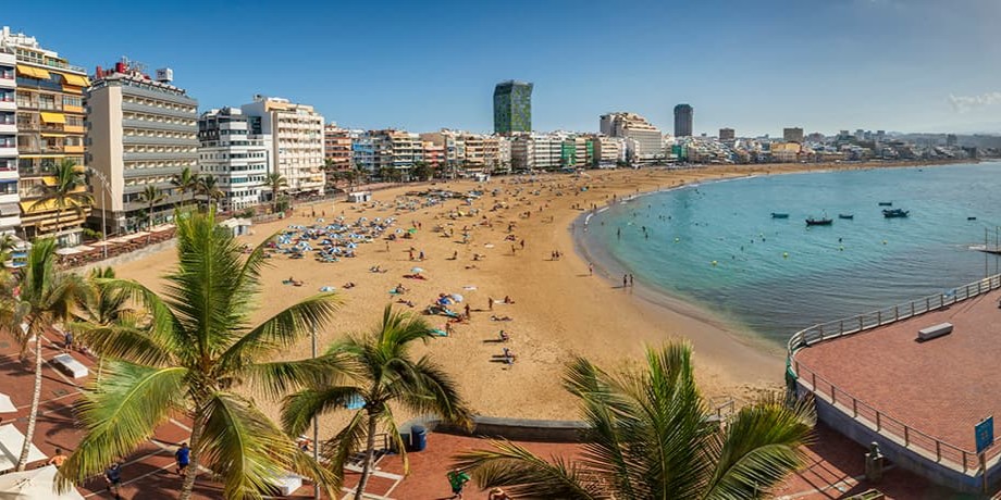Playa de las Canteras vista desde lo alto de un hotel, se puede ver el agua cristtalina, gente bañándose y en la arena tumbados, y al fondo varios hoteles costeros 