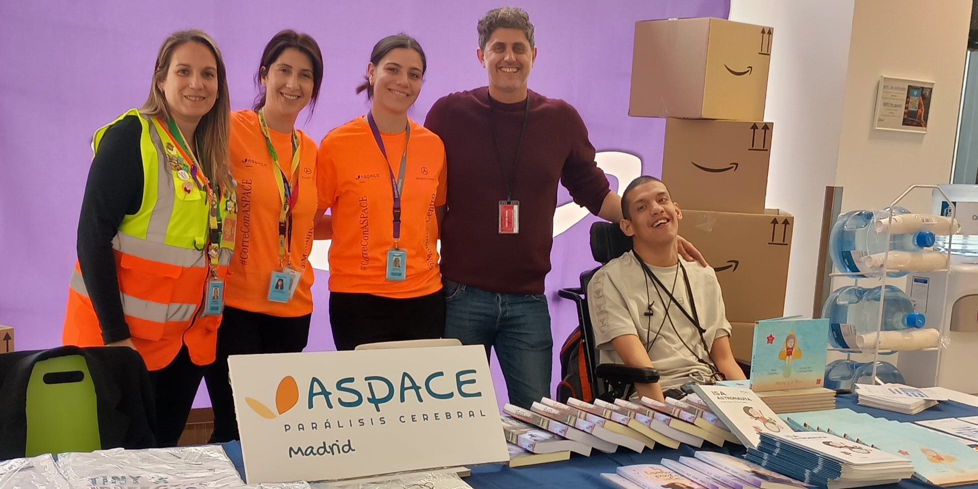 Stand de ASPACE Madrid en un espacio interior. Cinco personas posan juntas detrás de una mesa cubierta con materiales informativos, entre ellos libros, cuadernos y folletos. En la parte frontal destaca un cartel con el texto “ASPACE Parálisis Cerebral Madrid”. Tres participantes llevan camisetas naranjas con el nombre de la organización y acreditaciones colgadas al cuello. A la derecha hay varias cajas de cartón apiladas y un expositor con garrafas de agua. El fondo presenta una pared de color morado y una iluminación uniforme.
