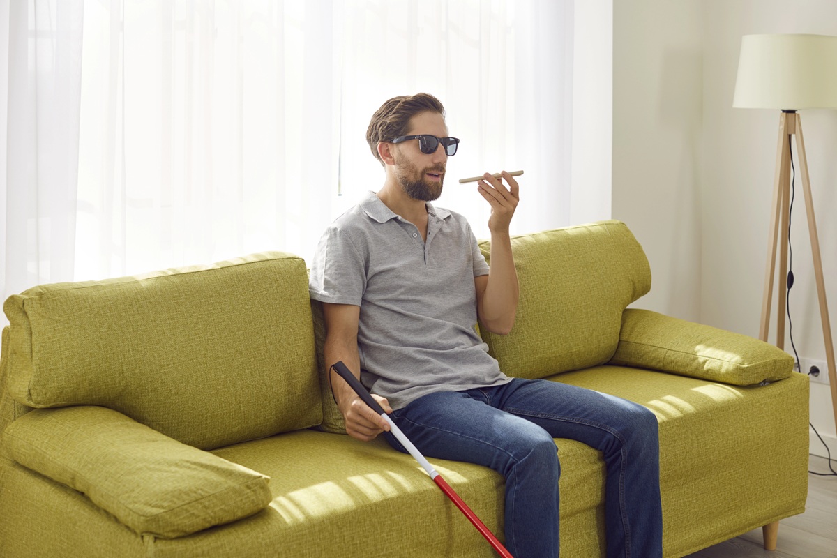 Hombre con discapacidad visual sentado en un sofá, usando gafas oscuras y sosteniendo un bastón blanco, mientras habla al teléfono móvil para realizar una tarea de accesibilidad digital.