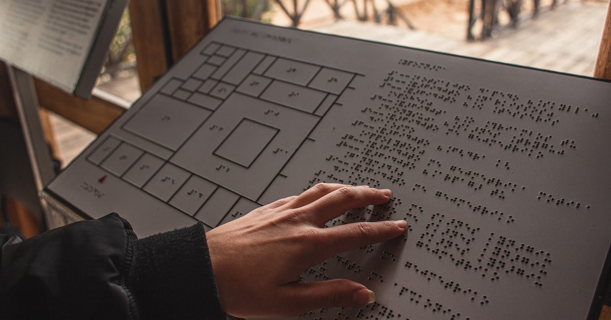 aparece la mano de una persona tocando una maqueta en la que aparece texto en braille