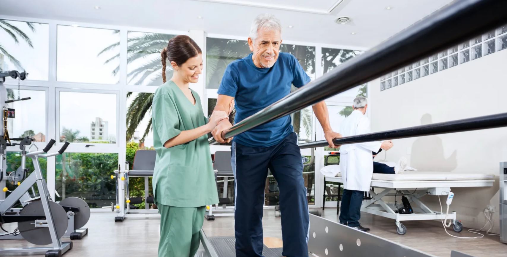 hombre agarrado a unas barras paralelas intentando andar con ayuda de una enfermera a su lado. Es una técnica rutinaria de los centros de dia