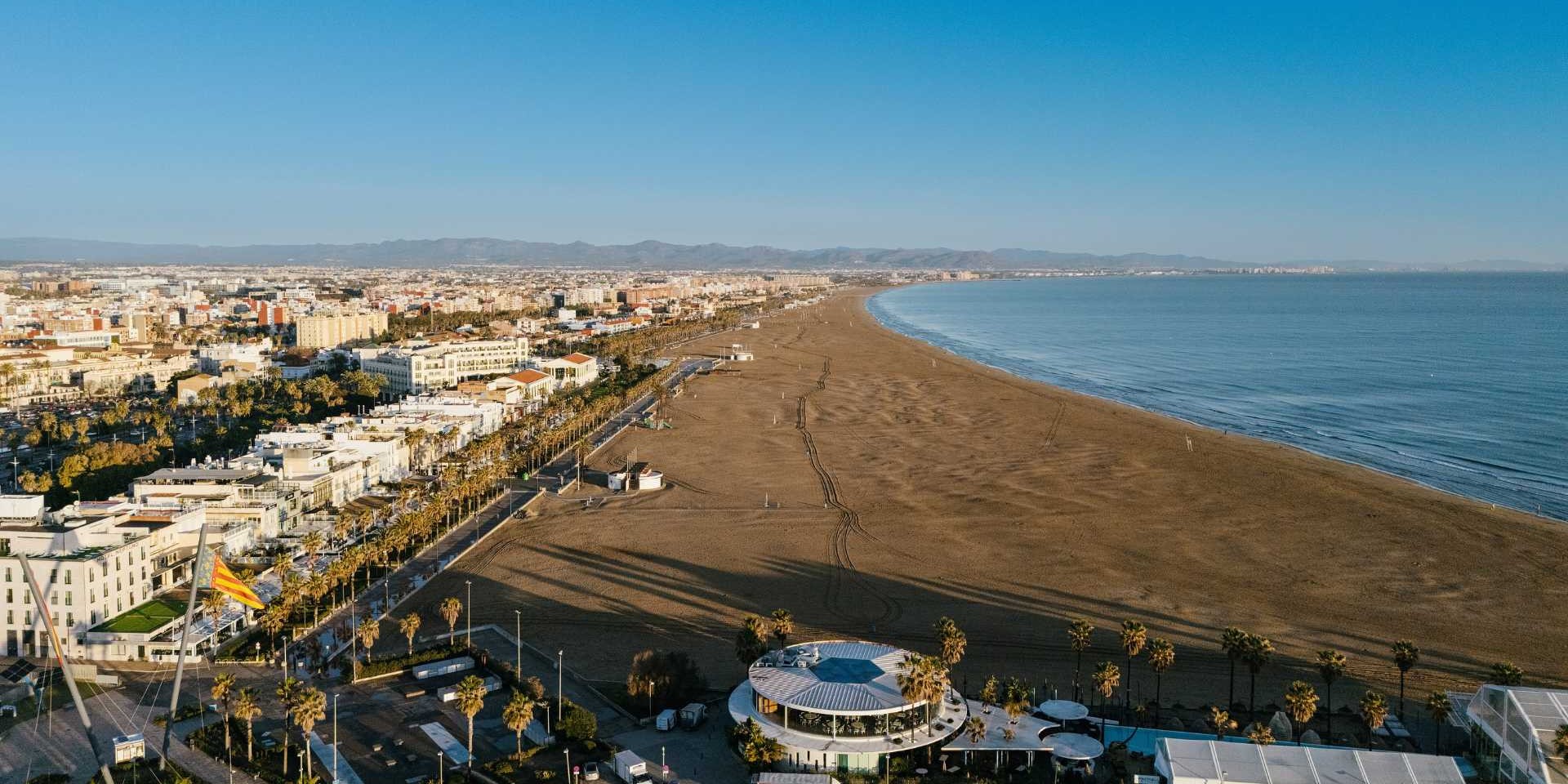 PLaya de la Malvarrosa con el mar a la derecha, en el centro una gran cala de arena fina y a la izquierda apartamentos y el paseo marítimo. Vista desde el aire.
