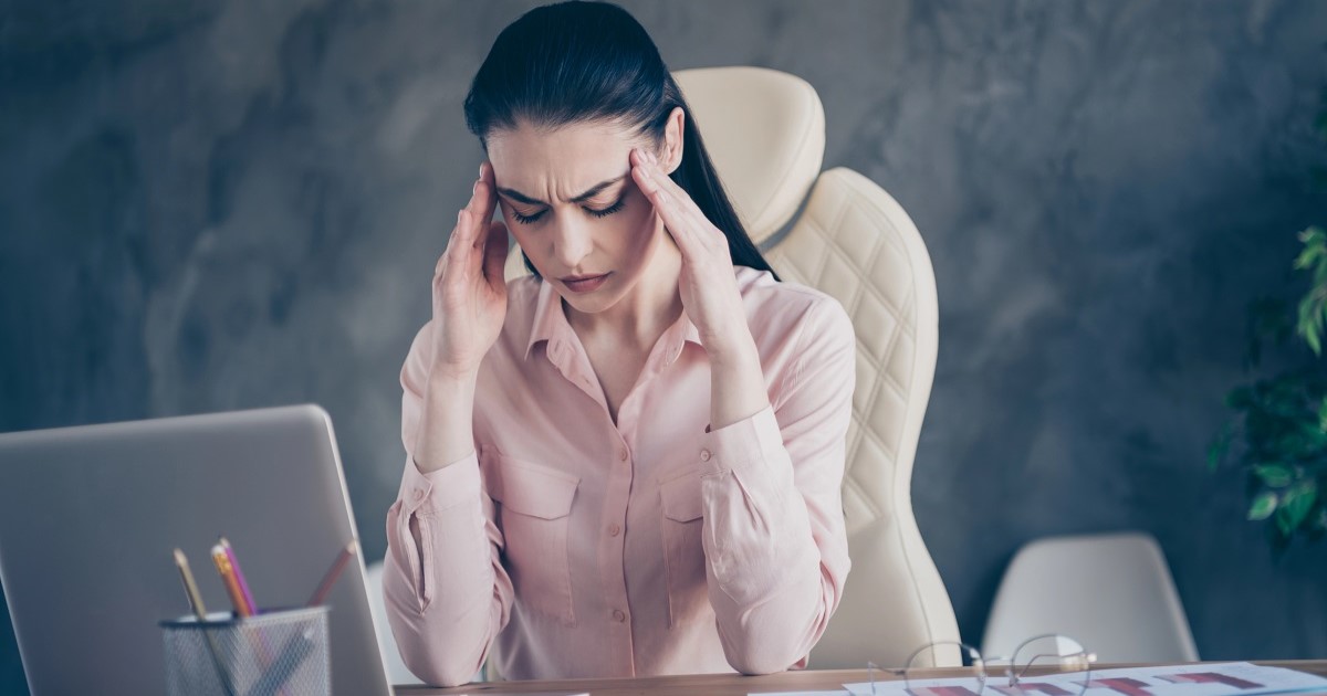mujer sentada en la silla de un escritorio con su portatil, en la mesa hay unas gráficas como si estuviese en su trabajo. La mujer se toca la cabeza como si la doliese.
