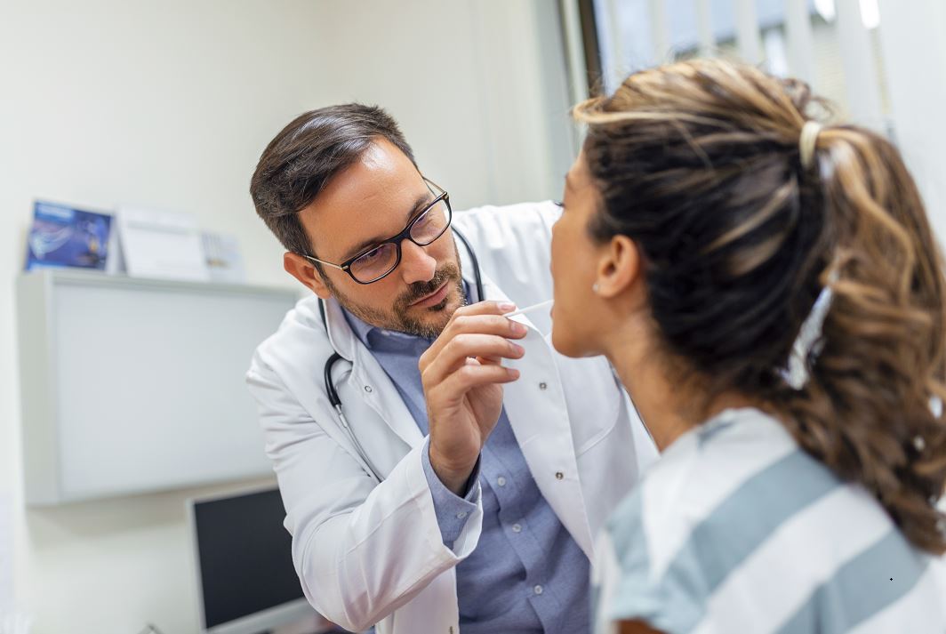 Una mujer está sentada frente a su médico. El médico se aproxima con un depresor de lengua mientras la mujer levanta la mirada y saca la lengua. Ejemplo de exámen médico ante una posible enfermedad infecciosa.