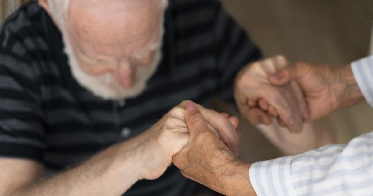 Una persona da la mano y ayuda a levantarse a una persona mayor con alzheimer, enfermedad neurodegenerativa.