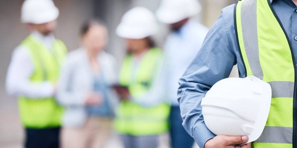 Trabajadores con chalecos reflectantes y cascos de seguridad en un entorno de obra; en primer plano, una persona sostiene un casco blanco mientras en el fondo otras personas consultan documentos o dispositivos, desenfocadas.