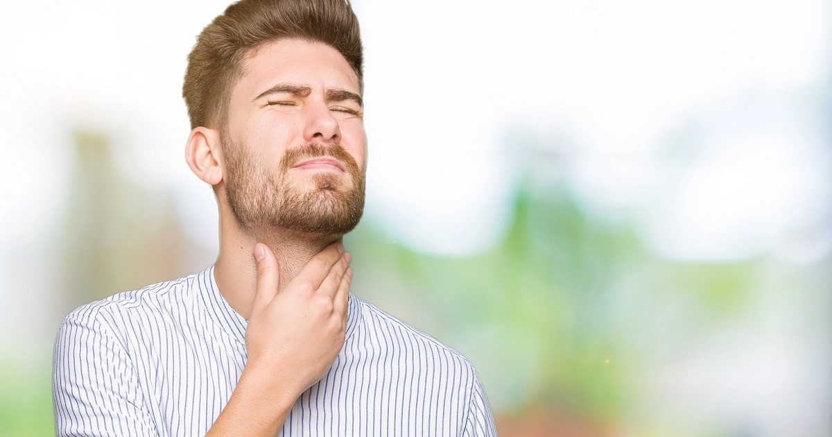 Un hombre con barba y camisa a rayas se toca el cuello con expresión de molestia. Su rostro refleja incomodidad, probablemente por picor de garganta. El fondo es borroso con tonos verdes y blancos, sugiriendo un ambiente al aire libre o bien iluminado.