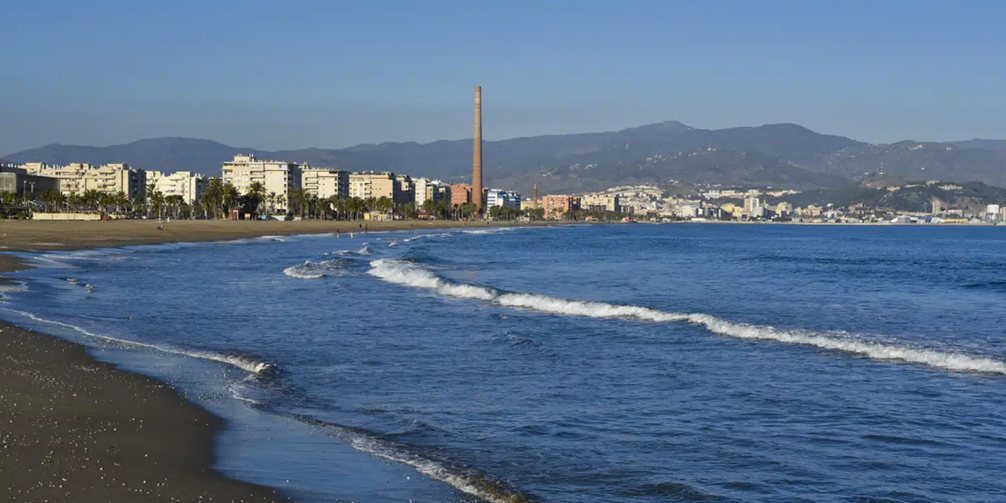 PLaya de la Misericordia con una imagen hecha desde la playa, al fondo se pueden ver apartamentos y hoteles, a la derecha la playa con olas y a la izquierda arena. Hay unas pocas personas bañándose