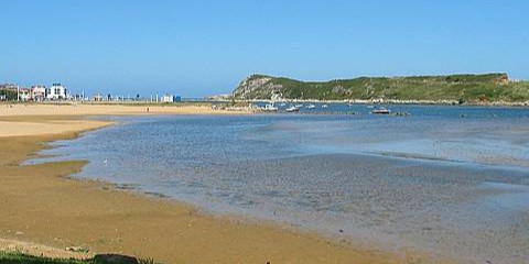 Playa de la Riberuca muy llana y de arena fina con un pequeño acantilado con vegetación al fondo al fondo 