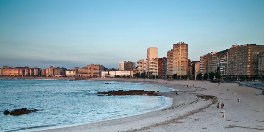 Playa de Riazor en un amanecer, con edificios al fondo y la playa a la izquierda. Se puede ver a gente paseando por la orilla