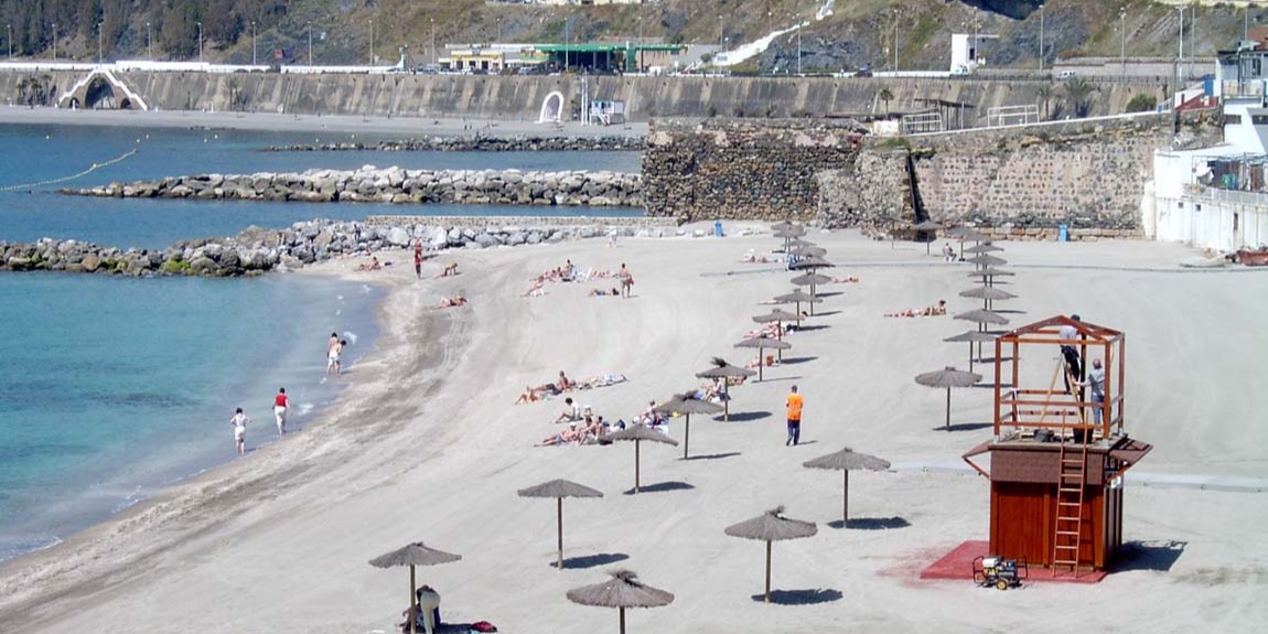 Foto completa de la playa de la Ribera, se ve a gente bañándose y otras tumbadas en la arena con sombrillas. También el puesto del socorrista y una llanura de fondo