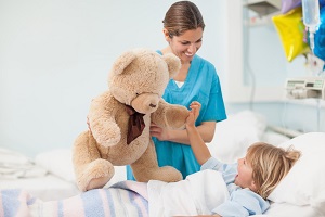 niño en cama de hospital con visita de una enfermera con un osito de peluche