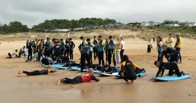 Participantes y monitores de una actividad de surf adaptado reciben formación en la playa de Loredo durante el I Festival de Parasurf Fundación También–Plea. Varias personas practican la posición sobre tablas de surf en la arena mientras un grupo observa y se prepara para entrar al agua. La imagen muestra una jornada de deporte inclusivo y aprendizaje junto al mar Cantábrico.