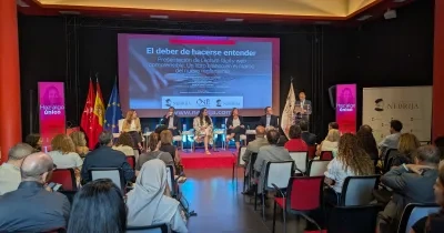 Público asistente y participantes en la presentación del libro Lectura Fácil y web comprensible. Un libro blanco en el marco del nuevo reglamento, celebrada en el Salón de Grados del Campus de Princesa de la Universidad Nebrija. En el escenario, varias personas intervienen en una mesa redonda mientras una persona realiza una presentación desde un atril. Al fondo se proyecta la imagen del evento y se muestran banderas institucionales.