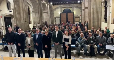 Salón de actos instalado en el interior de un edificio histórico con altos arcos y columnas de piedra. En primer plano, seis personas permanecen de pie frente a una mesa con micrófonos, documentos y botellas de agua. Detrás, decenas de asistentes ocupan las filas de asientos durante un acto público. La estancia está iluminada por luz artificial y por un gran ventanal circular situado en la parte superior del fondo de la sala. El público y las personas situadas en la mesa se distribuyen bajo una arquitectura