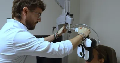 Profesional sanitario realiza una prueba de visión a una persona sentada utilizando un equipo optométrico en una consulta. La persona examinada lleva colocado un dispositivo frente a los ojos mientras el especialista ajusta el aparato para evaluar su capacidad visual. El entorno es una sala clínica con equipamiento oftalmológico.