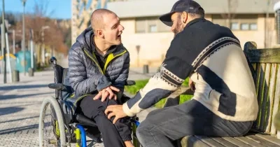 Dos hombres, un de ellos en silla de ruedas y el otro sentado en un banco, hablando