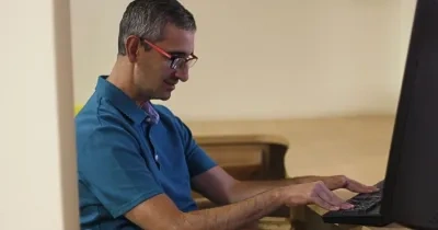 Persona sentada frente a un ordenador de sobremesa, utilizando el teclado en un entorno de trabajo interior. La imagen muestra un puesto de oficina con una pantalla, una mesa de madera y una iluminación suave.