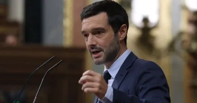 Persona con traje oscuro interviene desde un atril con dos micrófonos en el interior de una cámara parlamentaria. La imagen muestra a la persona realizando un gesto con la mano mientras se dirige al público, con el fondo desenfocado y elementos arquitectónicos de la sala visibles.