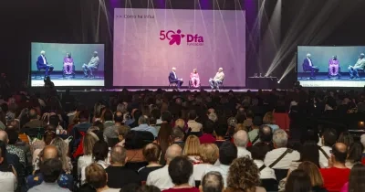 Dos personas subidas en el escenario hablando sentados en dos sillas, mientras hay multitud de personas en la grada observando 