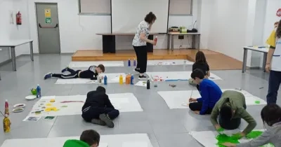 Alt text:  Grupo de menores participa en una actividad artística inclusiva en una sala escolar, pintando sobre grandes hojas de papel colocadas en el suelo. Varias personas trabajan con pinceles y pintura de colores mientras una profesional supervisa la actividad. La imagen refleja un entorno educativo colaborativo orientado al aprendizaje y la participación.