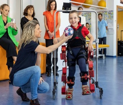Una mujer entrega un balón a un menor de la Fundación Bobath, miembro de Aspace que utiliza un exoesqueleto pediátrico de pie (Fuente: Marsi Bionics)