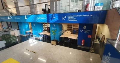 Vista interior de la Oficina de Información y Registro del Paseo del Prado, con varios puestos numerados (6, 7 y 8) señalizados con grandes paneles azules. Cada puesto incluye pictogramas, colores diferenciados y símbolos de accesibilidad. Se observan mesas, sillas, estanterías y un ascensor cercano. La señalética es clara y de alto contraste para facilitar la orientación.