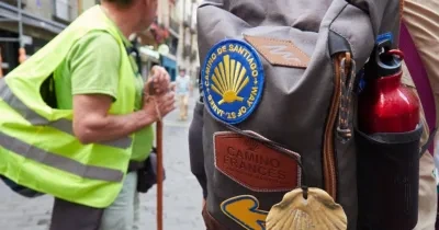 Un peregrino de fondo con un chaleco reflectante y a primera vista de la imagen una mochila negra con un parche del Camino de Santiago Francés, una cantimplora y una concha