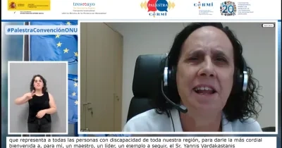 Captura de una videoconferencia institucional. En el centro, una persona sentada en un despacho, frente a la cámara, con auriculares y micrófono, interviene desde su puesto de trabajo. A la izquierda de la pantalla aparece una intérprete de lengua de signos española sobre fondo neutro. En el margen superior se muestran los logotipos del Gobierno de España, Fundación CERMI Mujeres y otras entidades, junto al texto “#PalestraConvenciónONU”. En la parte inferior se ven subtítulos en español.