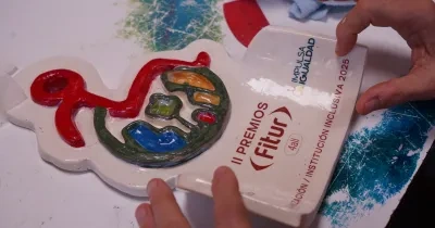 Trofeo de cerámica roto con diseño colorido y texto de los "II PREMIOS Fitur 4all 2025". Manos lo sostienen sobre una mesa con pintura, sugiriendo reparación o restauración.