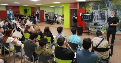 Grupo de personas asistiendo a una sesión formativa o conferencia en una sala accesible. Una persona realiza una presentación frente al público junto a una pantalla, mientras otra persona interpreta en lengua de signos. Entre los asistentes hay personas usuarias de silla de ruedas. La sala cuenta con paredes de colores vivos y un mural de cascadas al fondo.