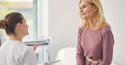 Persona sentada en una consulta médica conversa con un profesional sanitario. Ambos aparecen en una sala de exploración luminosa, con equipamiento clínico al fondo, en una escena relacionada con el asesoramiento y seguimiento de la salud durante la menopausia.