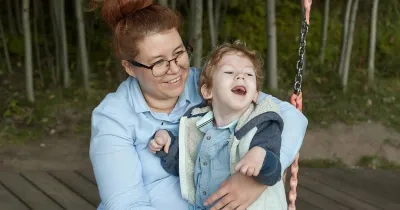 Persona adulta con gafas sostiene a un niño con discapacidad en un columpio adaptado al aire libre. Ambos se encuentran en una zona de juego rodeada de árboles. La imagen refleja acompañamiento, inclusión y apoyo a la infancia con discapacidad en un entorno accesible y seguro.
