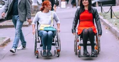 Dos personas usuarias de silla de ruedas se desplazan por una vía urbana peatonal. Una de ellas viste una camiseta clara y lleva un bolso cruzado, mientras que la otra viste un vestido rojo y una chaqueta oscura. A su alrededor caminan otras personas y se observan vehículos aparcados, árboles y mobiliario urbano al fondo. La escena transcurre al aire libre en un entorno urbano accesible.
