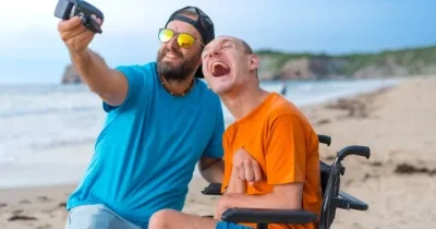 Una persona sin discapacidad junto a una persona con discapacidad en silla de ruedas haciéndose un selfie en la playa