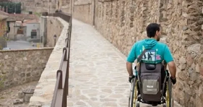 Una persona en silla de ruedas de espaldas transitando por las calles y la muralla de Ávila (Fuente: Ciudades Patrimonio)
