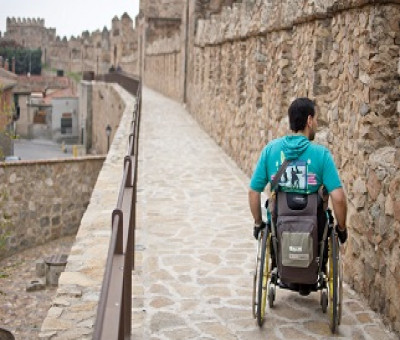 Una persona en silla de ruedas de espaldas transitando por las calles y la muralla de Ávila (Fuente: Ciudades Patrimonio)