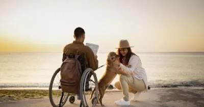 Una persona en silla de ruedas disfrutando de su tiempo de ocio turístico frente al mar, junto a su pareja y su perro (Fuente: Servimedia)