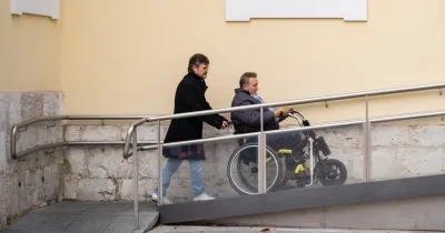 hombre ayudando a una persona en silla de ruedas subir por una rampa accediendo a un edificio.