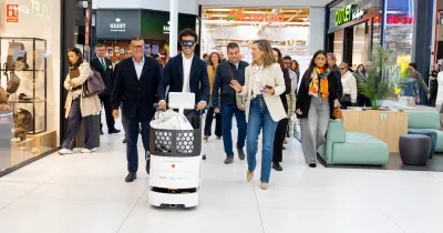Persona guiando un robot asistencial autónomo por el pasillo de un centro comercial. Varias personas acompañan la demostración y observan el recorrido del dispositivo, en un espacio comercial amplio y luminoso con tiendas y zonas de descanso al fondo. La imagen muestra una prueba de tecnología orientada a mejorar la accesibilidad y la movilidad en entornos comerciales.
