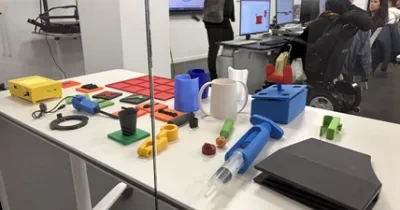 Mesa de trabajo con diversos objetos fabricados mediante impresión 3D, entre ellos recipientes, piezas de ensamblaje, una taza, adaptadores y herramientas de apoyo en distintos colores. Al fondo, tras un cristal decorado con triángulos rojos, varias personas participan en una actividad formativa frente a ordenadores mientras observan diseños digitales mostrados en las pantallas.