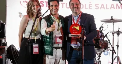 Tres personas posan sobre un escenario durante un acto de Startup Olé Salamanca. Una de ellas sostiene un premio de color rojo mientras otra hace el gesto de aprobación con el pulgar. En primer plano hay varios micrófonos y, al fondo, se aprecia una batería y el logotipo del evento proyectado en una pantalla.