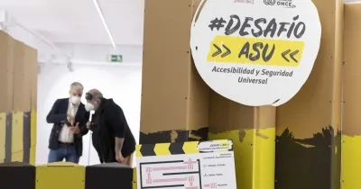 Espacio interior con paneles altos en colores amarillo, negro y marrón que forman un recorrido del proyecto “#Desafío ASU – Accesibilidad y Seguridad Universal”. En primer plano se ve un cartel circular con el nombre del proyecto y, detrás, dos personas observando una estructura del circuito. La escena muestra un entorno expositivo dedicado a la accesibilidad.